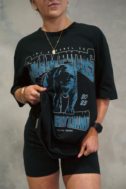 Female wearing TMJ Apparel 1993 Champions Tee in Blue with a large print on the front with the words “TMJ Apparel Co”, “Champions” “Earn Everything” &amp; Premium Athleticulture Goods”.