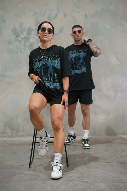 Male &amp; Female wearing TMJ Apparel 1993 Champions Tee in Blue with a large print on the front with the words “TMJ Apparel Co”, “Champions” “Earn Everything” &amp; Premium Athleticulture Goods”.