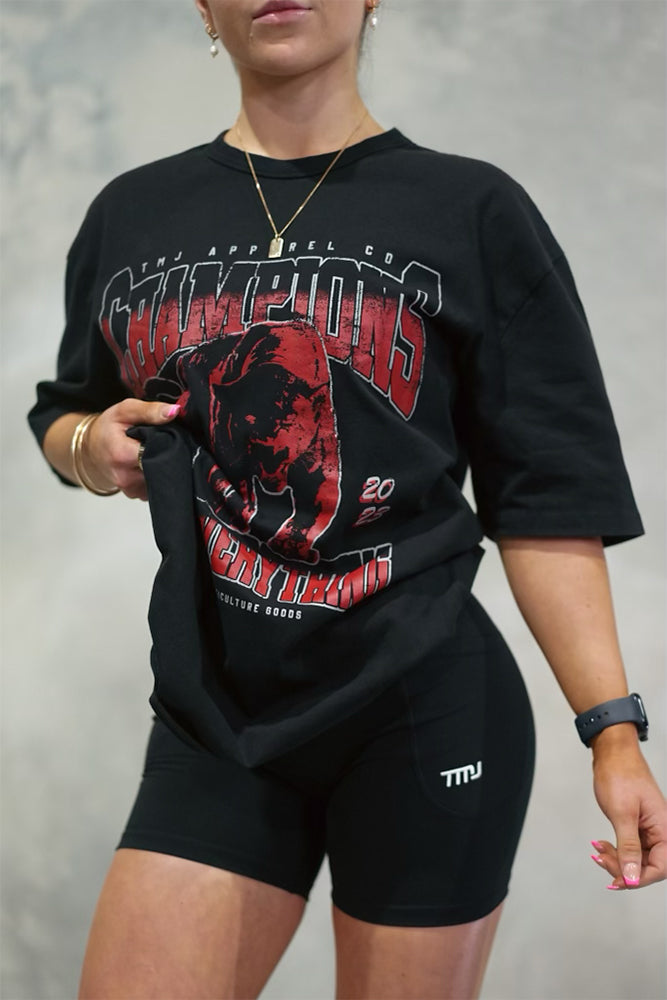 Female wearing TMJ Apparel 1993 Champions Tee in Red with a large print on the front with the words “TMJ Apparel Co”, “Champions” “Earn Everything” &amp; Premium Athleticulture Goods”.