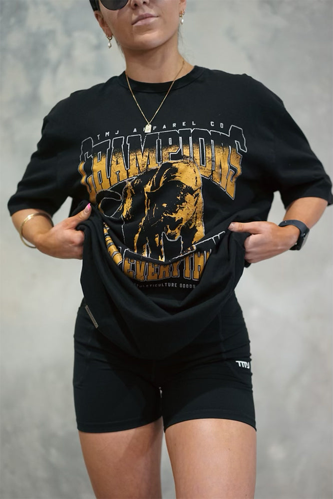 Female wearing TMJ Apparel 1993 Champions Tee in Gold with a large print on the front with the words “TMJ Apparel Co”, “Champions” “Earn Everything” &amp; Premium Athleticulture Goods”.