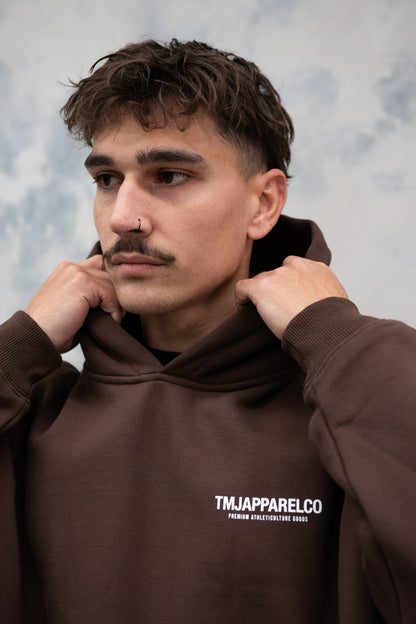 Male wearing TMJ Apparel Athleticulture Hoodie in Brown showing the front of the hoodie with white text saying TMJAPPARELCO Premium Athleticulture Goods on left of chest.