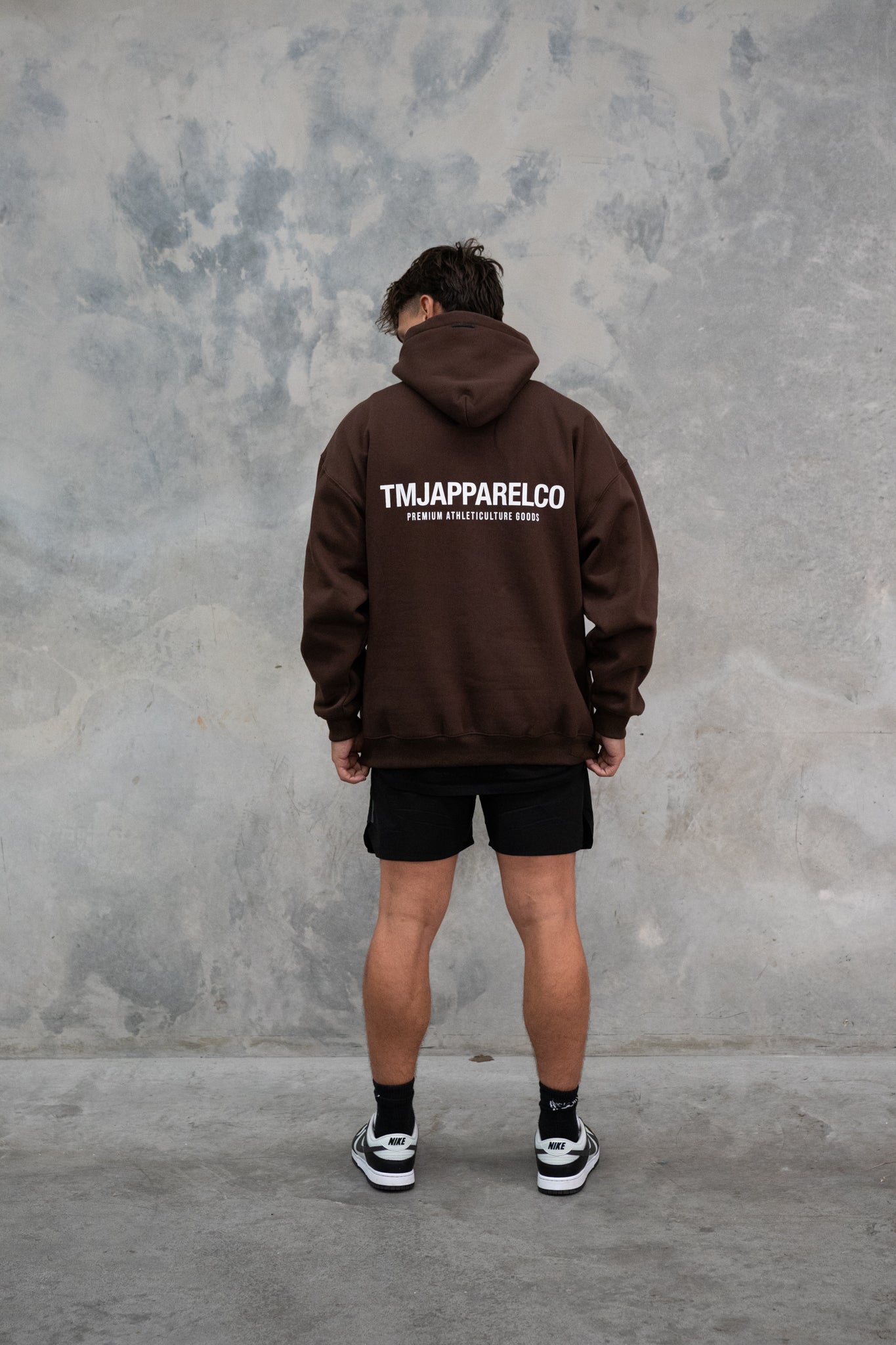  Male wearing TMJ Apparel Athleticulture Hoodie in Brown showing the back of the hoodie with white text saying TMJAPPARELCO Premium Athleticulture Goods in the middle centre of back.