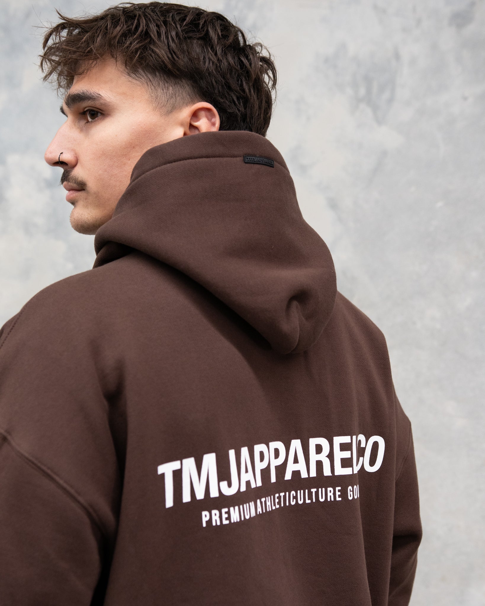  Male wearing TMJ Apparel Athleticulture Hoodie in Brown showing the back of the hoodie with white text saying TMJAPPARELCO Premium Athleticulture Goods in the middle centre of back.