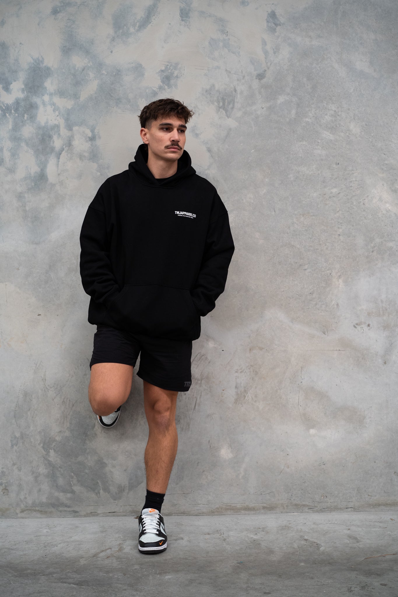 Male wearing TMJ Apparel Athleticulture Hoodie in Black showing the front of the hoodie with white text saying TMJAPPARELCO Premium Athleticulture Goods on left of chest.