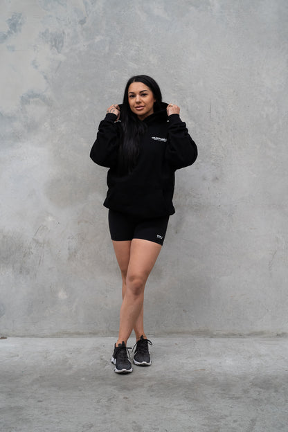 Female wearing TMJ Apparel Athleticulture Hoodie in Black showing the front of the hoodie with white text saying TMJAPPARELCO Premium Athleticulture Goods on left of chest.