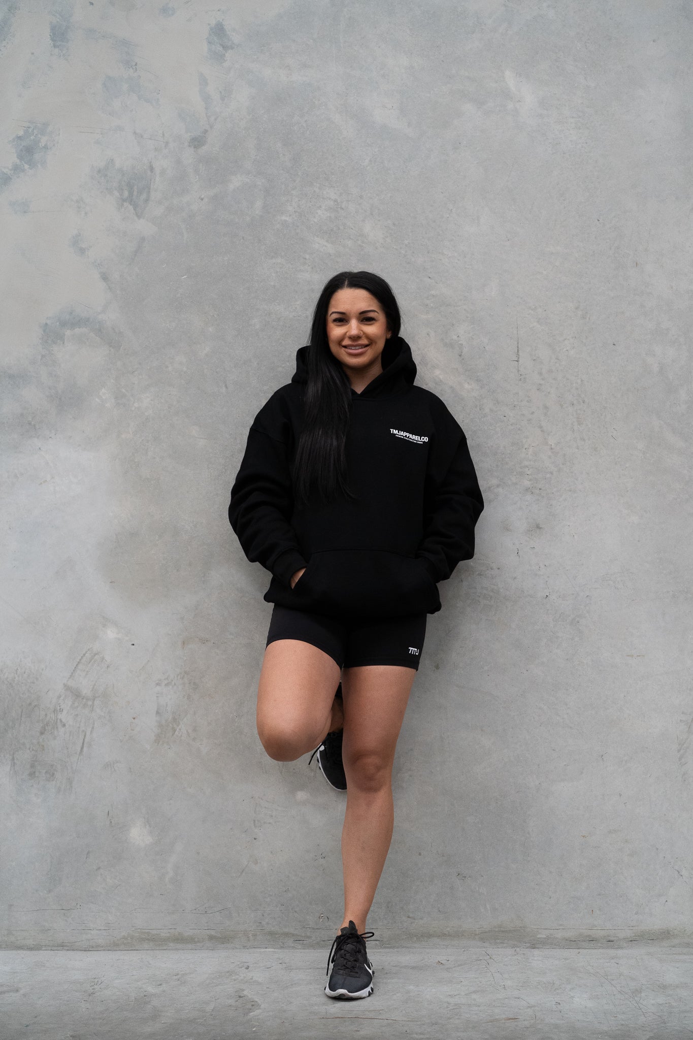 Female wearing TMJ Apparel Athleticulture Hoodie in Black showing the front of the hoodie with white text saying TMJAPPARELCO Premium Athleticulture Goods on left of chest.