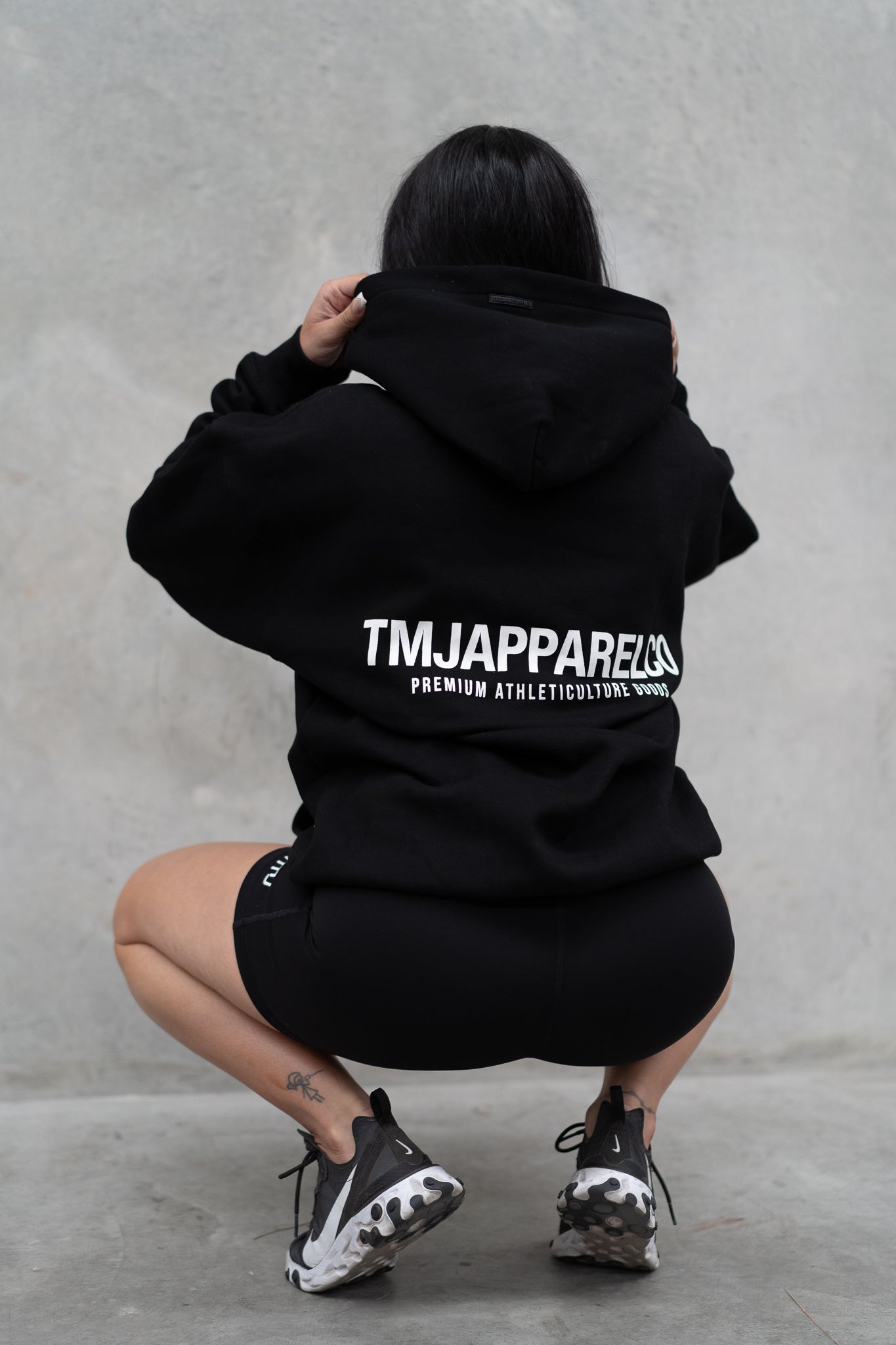 Female wearing TMJ Apparel Athleticulture Hoodie in Black showing the back of the hoodie with white text saying TMJAPPARELCO Premium Athleticulture Goods in the middle centre of back.