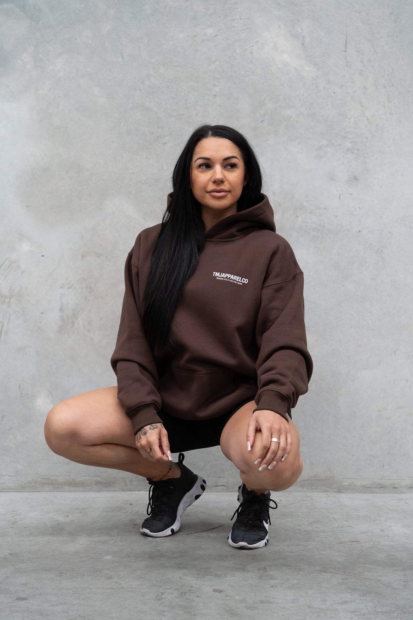 Female wearing TMJ Apparel Athleticulture Hoodie in Brown showing the front of the hoodie with white text saying TMJAPPARELCO Premium Athleticulture Goods on left of chest.