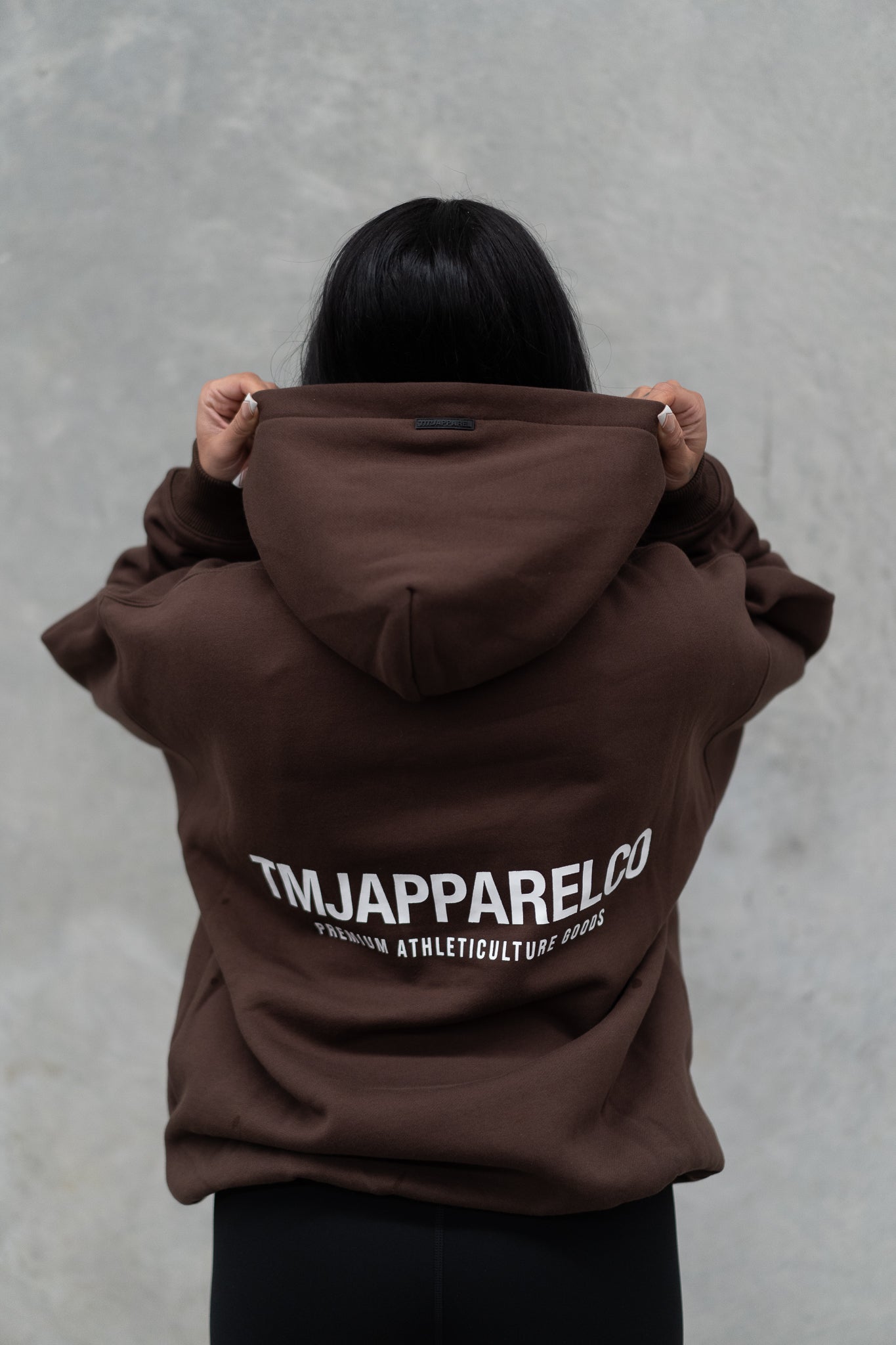Female wearing TMJ Apparel Athleticulture Hoodie in Brown showing the back of the hoodie with white text saying TMJAPPARELCO Premium Athleticulture Goods in the middle centre of back.