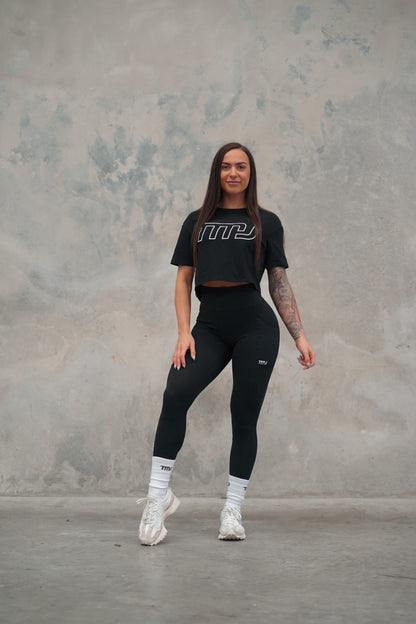 Female wearing TMJ Apparel Cropped Tee in Black showing the large white “TMJ” outline logo across the chest.