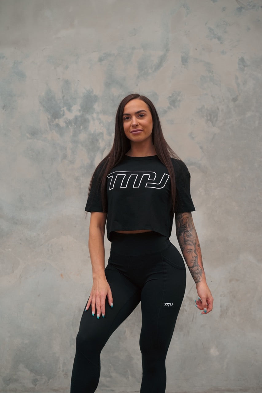 Female wearing TMJ Apparel Cropped Tee in Black showing the large white “TMJ” outline logo across the chest.