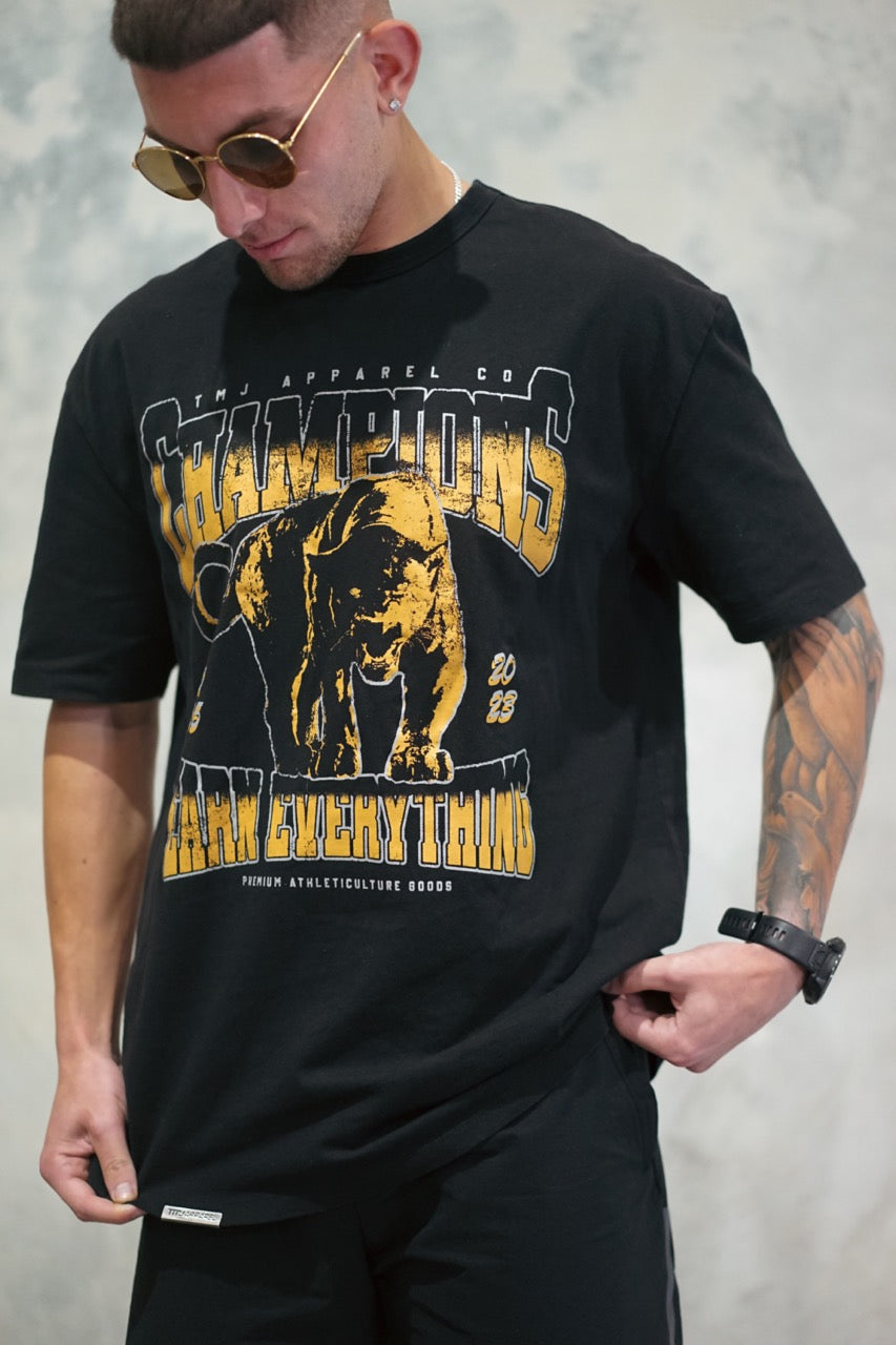 Male wearing TMJ Apparel 1993 Champions Tee in Gold with a large print on the front with the words “TMJ Apparel Co”, “Champions” “Earn Everything” &amp; Premium Athleticulture Goods”.