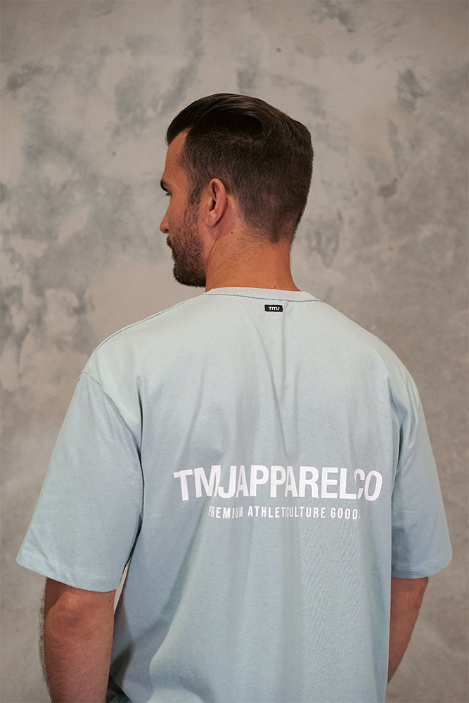 Male wearing TMJ Apparel Athleticulture Tee in Powder Blue showing the back of the shirt with white text saying TMJAPPARELCO Premium Athleticulture Goods in the the middle centre of back