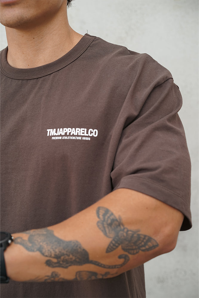 Male wearing TMJ Apparel Athleticulture Tee in Brown front of the shirt with white text saying TMJAPPARELCO Premium Athleticulture Goods on left of chest