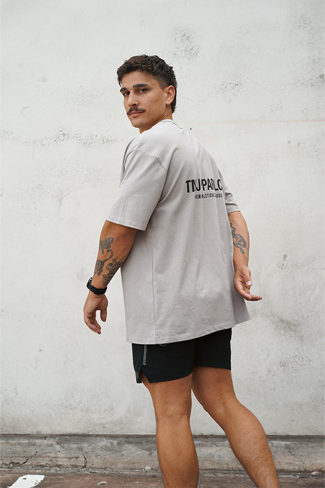 Male wearing TMJ Apparel Athleticulture Tee in Dune showing the back of the shirt with white text saying TMJAPPARELCO Premium Athleticulture Goods in the the middle centre of back