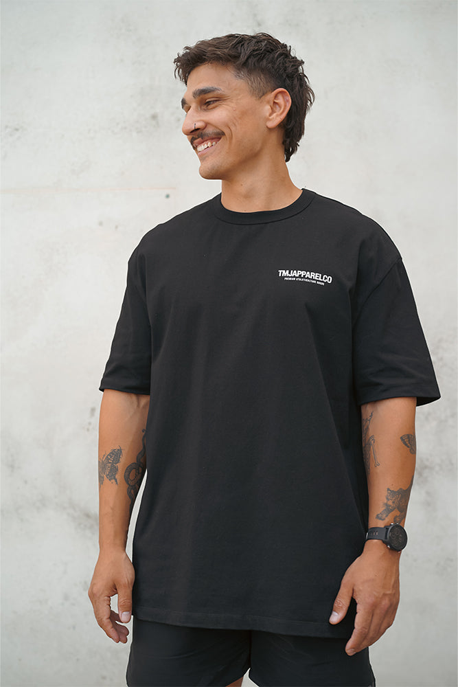 Male wearing TMJ Apparel Athleticulture Tee in Black showing the front of the shirt with white text saying TMJAPPARELCO Premium Athleticulture Goods on left of chest